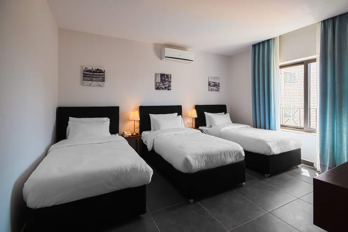 Quarto de hotel com três camas, uma janela com cortinas azuis e dois aparelhos de ar condicionado fixados na parede.