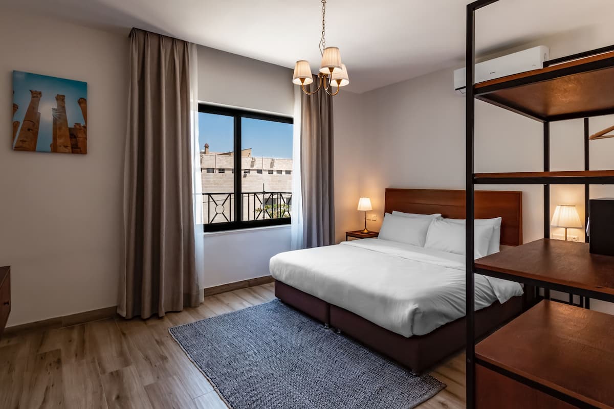 Quarto de hotel com uma cama grande, uma escrivaninha, uma janela com cortinas, um candelabro e um quadro na parede.