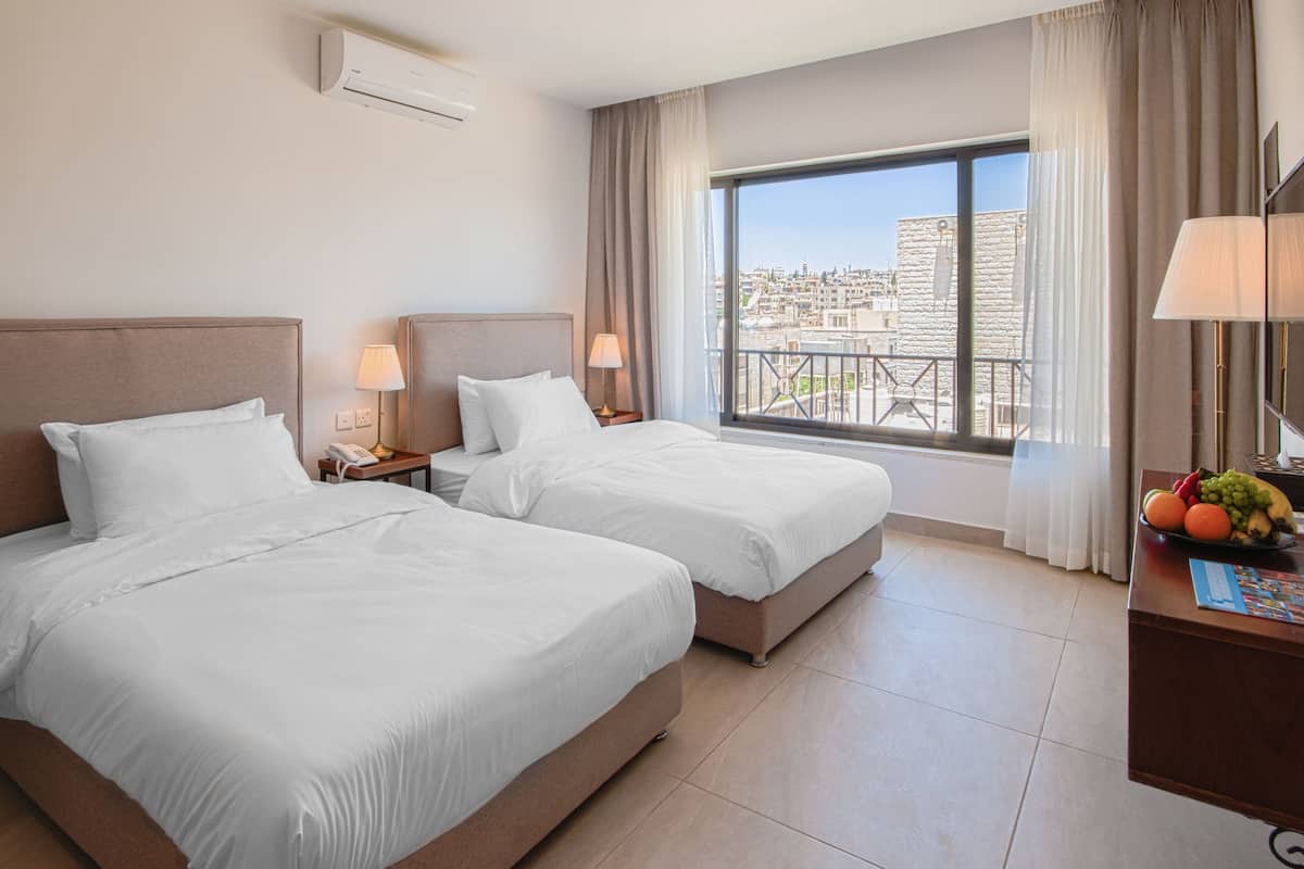 Quarto de hotel com duas camas, varanda com vista para a cidade, escrivaninha com luminária e uma cesta de frutas.