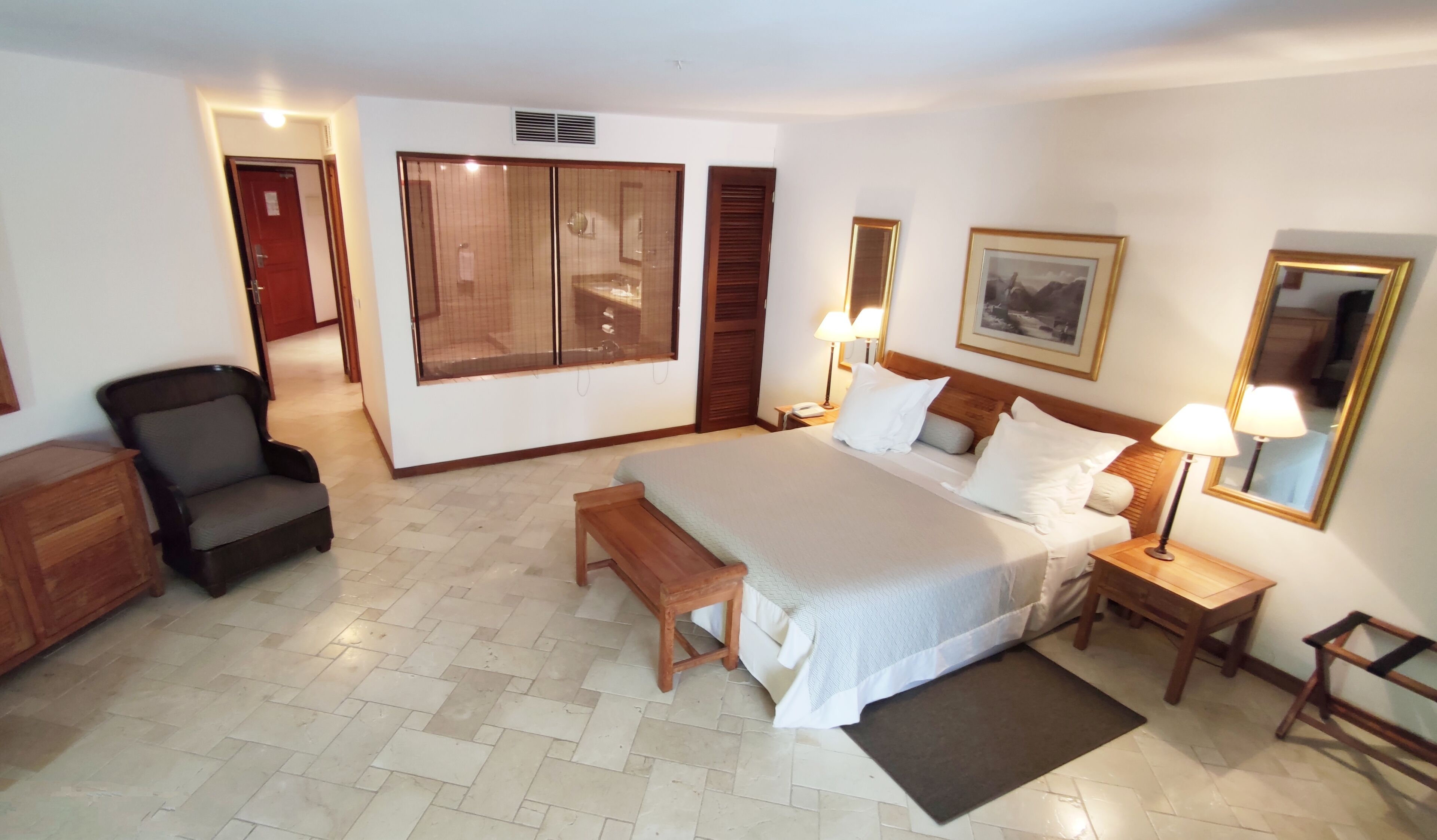 A hotel room with a bed, bedside tables, a chair, a desk, a wardrobe, and a large window.