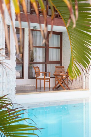 Zona de piscina con dos sillas y una mesa de madera, una ventana con cortinas y vista al estanque.