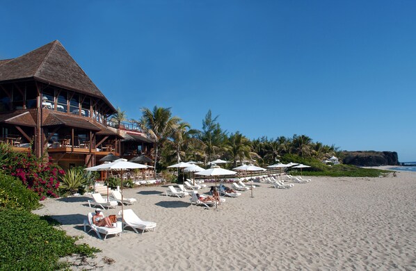 Un hotel frente a la playa con techo de paja, varias zonas de estar con sombrillas y vistas al océano.