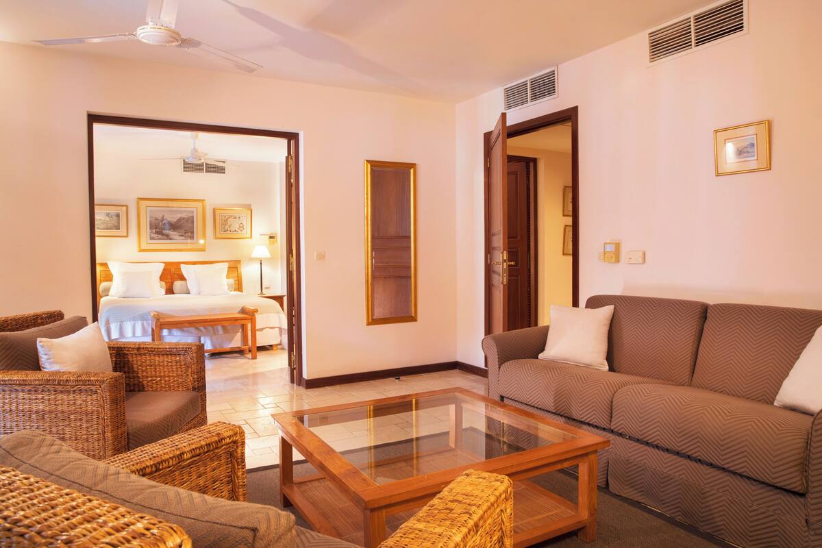 Una habitación de hotel con cama, sofá, mobiliario de mimbre, una mesa de centro de vidrio y cuadros en las paredes.