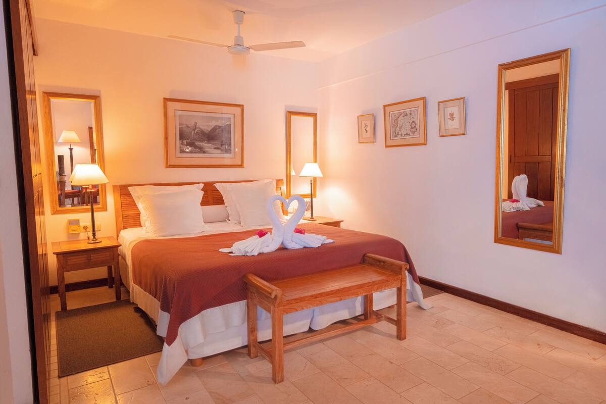 Habitación de hotel con una cama grande, dos mesitas de noche con lámparas, un banco de madera y un ventilador de techo.