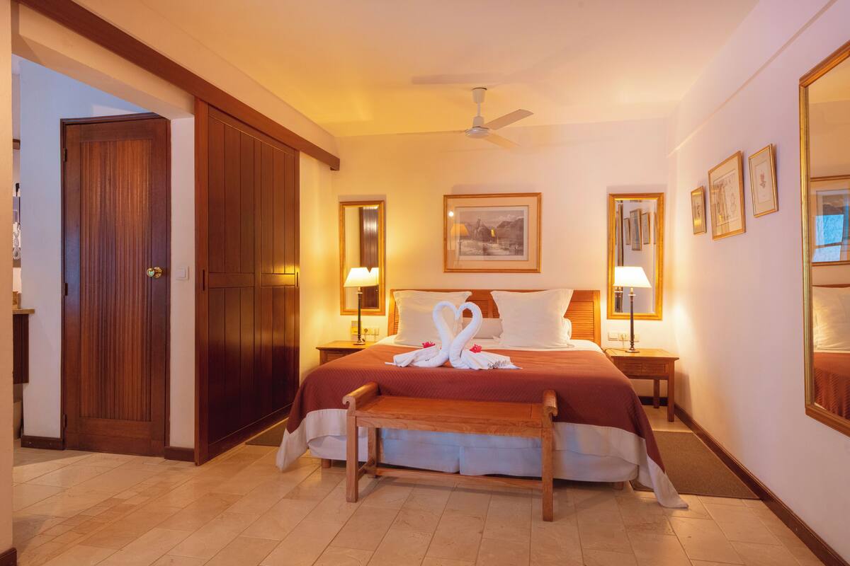 Habitación de hotel con una cama grande, dos lámparas de noche, un ventilador de techo, un banco de madera y un espejo grande en la pared.