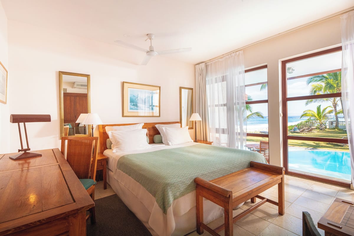 Habitación de hotel con cama, escritorio, silla, mesa, ventilador de techo y vista a una piscina y palmeras.