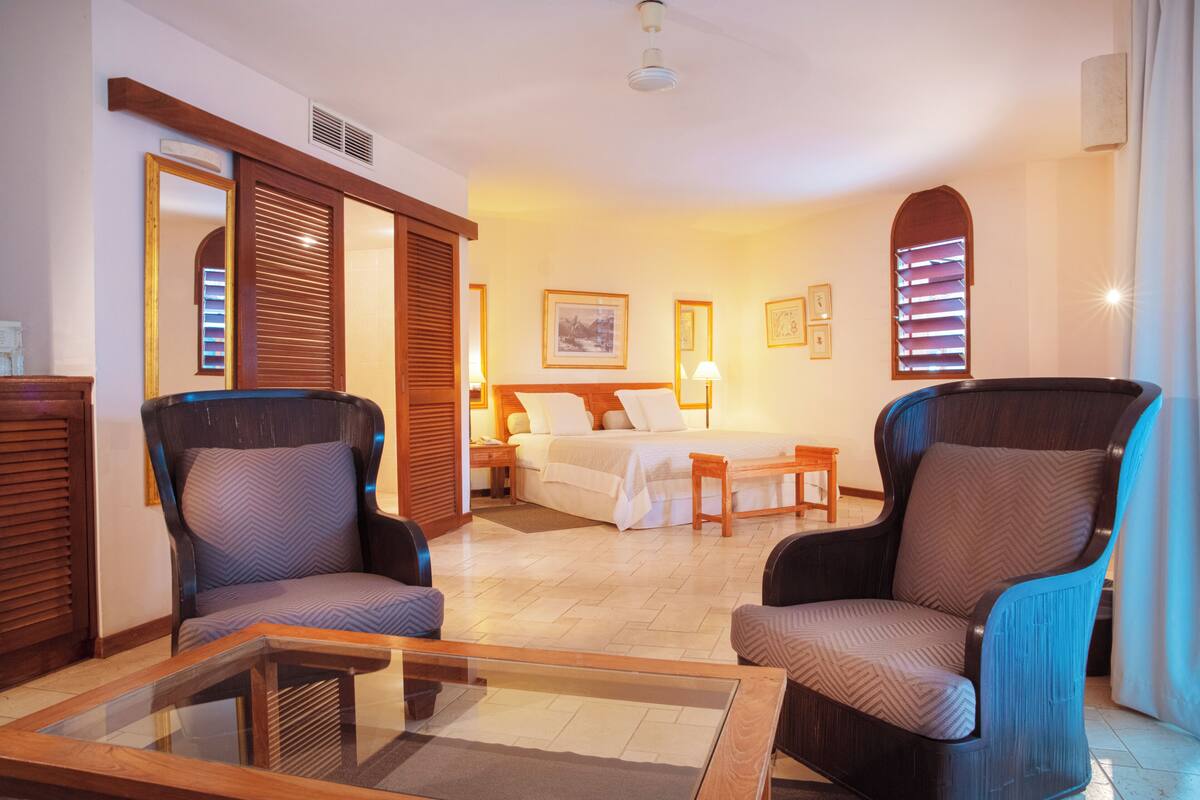 Habitación de hotel con una cama, dos sillones, una mesa de centro de vidrio, una mesita de noche de madera y un espejo grande.
