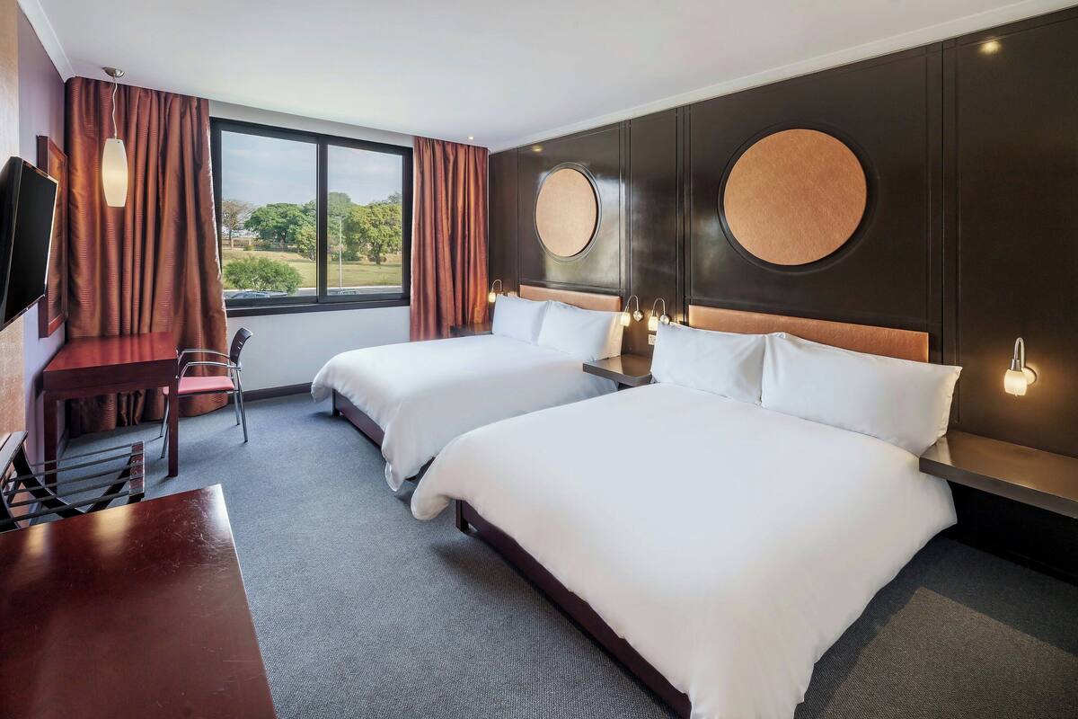 Une chambre d’hôtel avec deux lits, un bureau et une chaise. Elle dispose d’une grande fenêtre avec des rideaux, d’une télévision et de lampes fixées au mur.