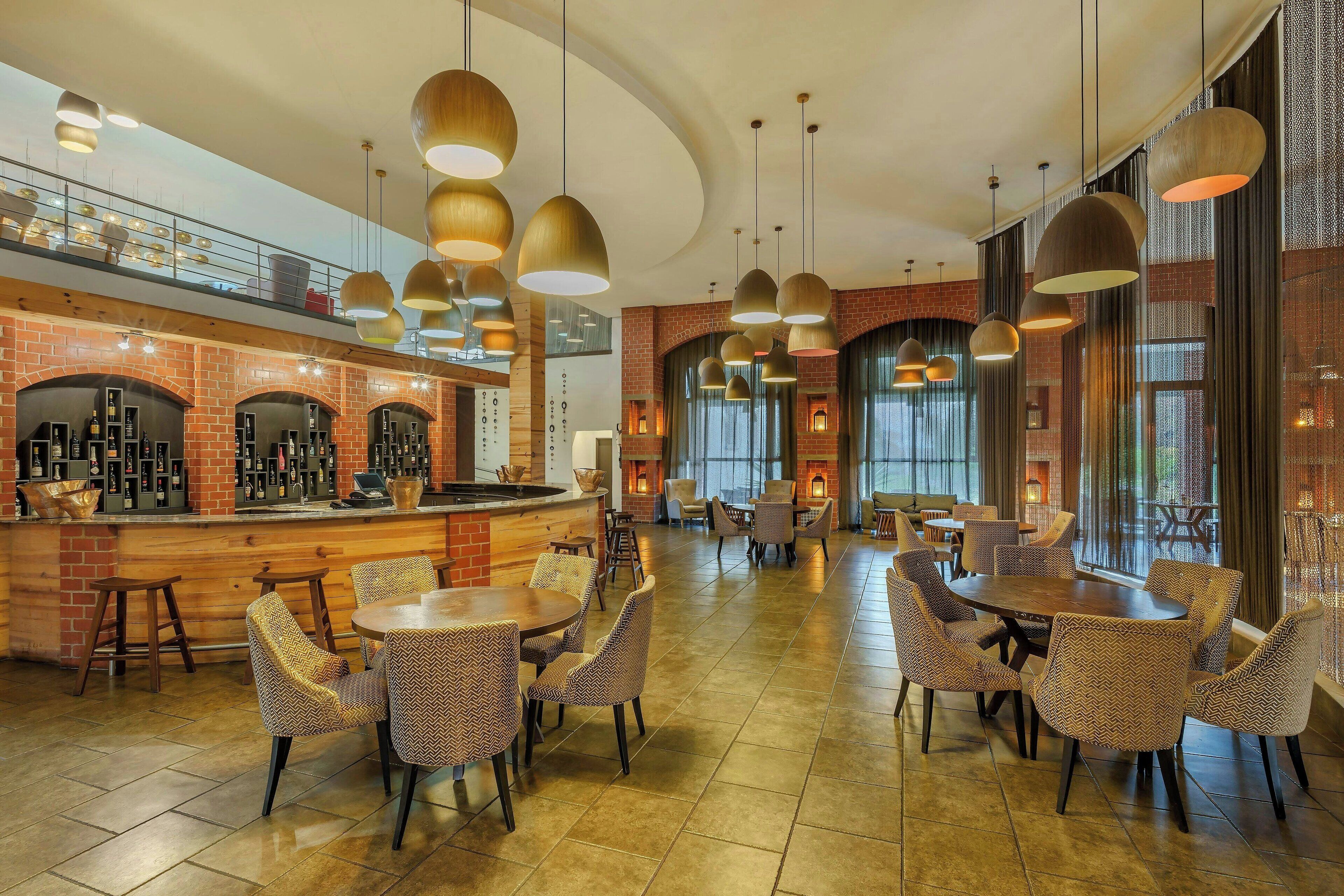 A spacious restaurant with high ceilings, hanging pendant lights, brick walls, and a variety of seating arrangements including round tables and chairs.