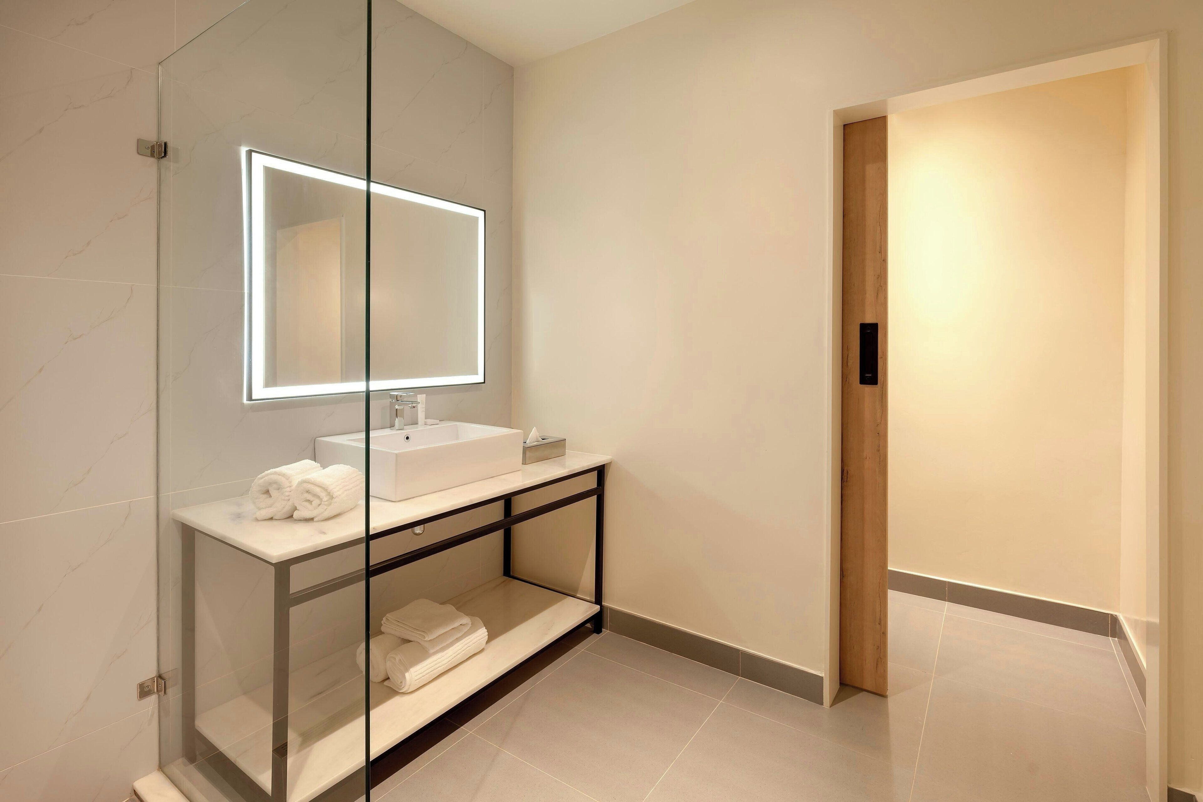 A modern bathroom with a glass-enclosed vanity, a large mirror with integrated lighting, and a wooden door.