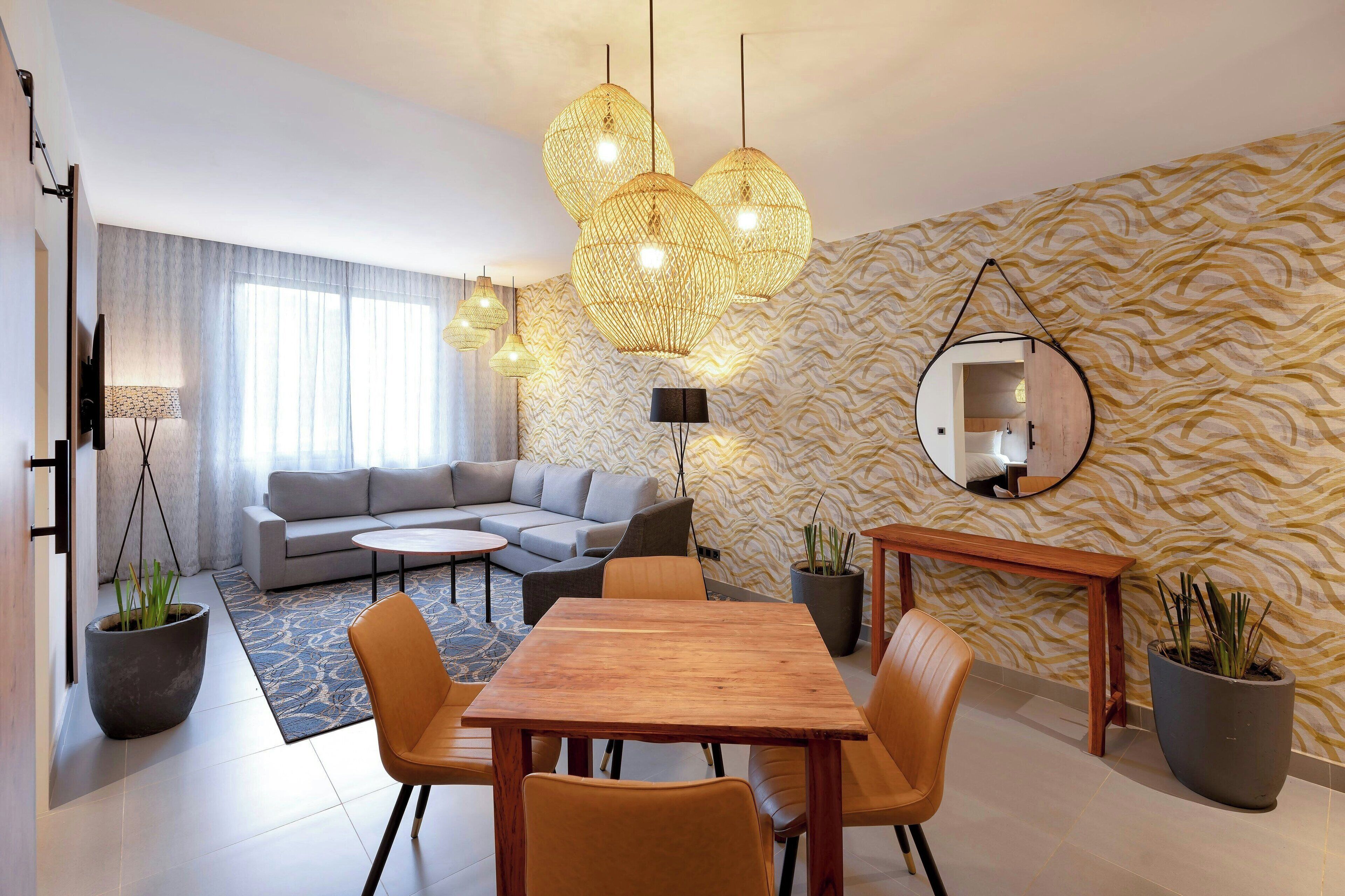 A modern living room with a sofa, a round coffee table, a wooden dining table with chairs, hanging wicker lamps, a large mirror, and patterned wallpaper.