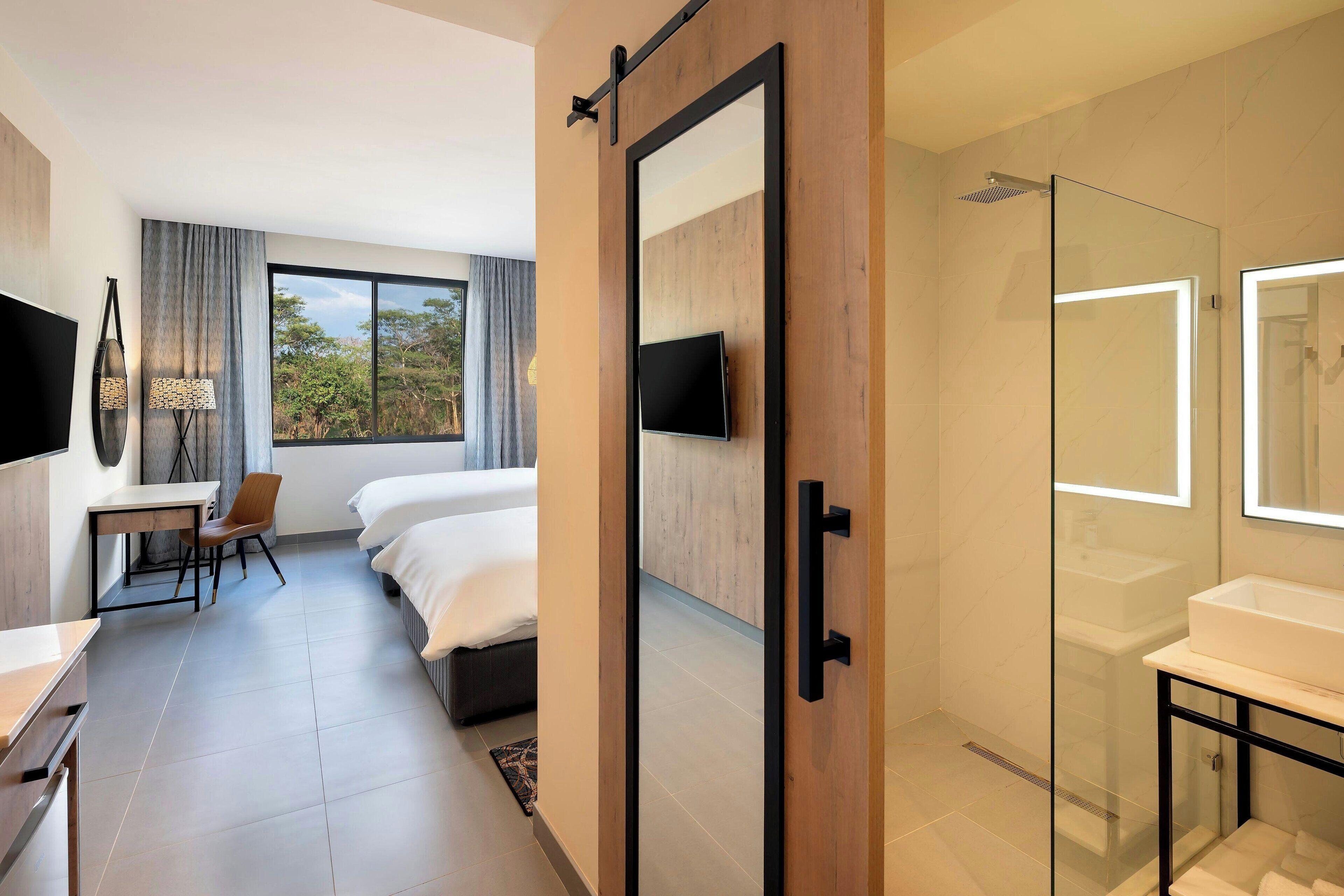 A modern hotel room with a bed, a desk, a TV, a shower area, and a large window.