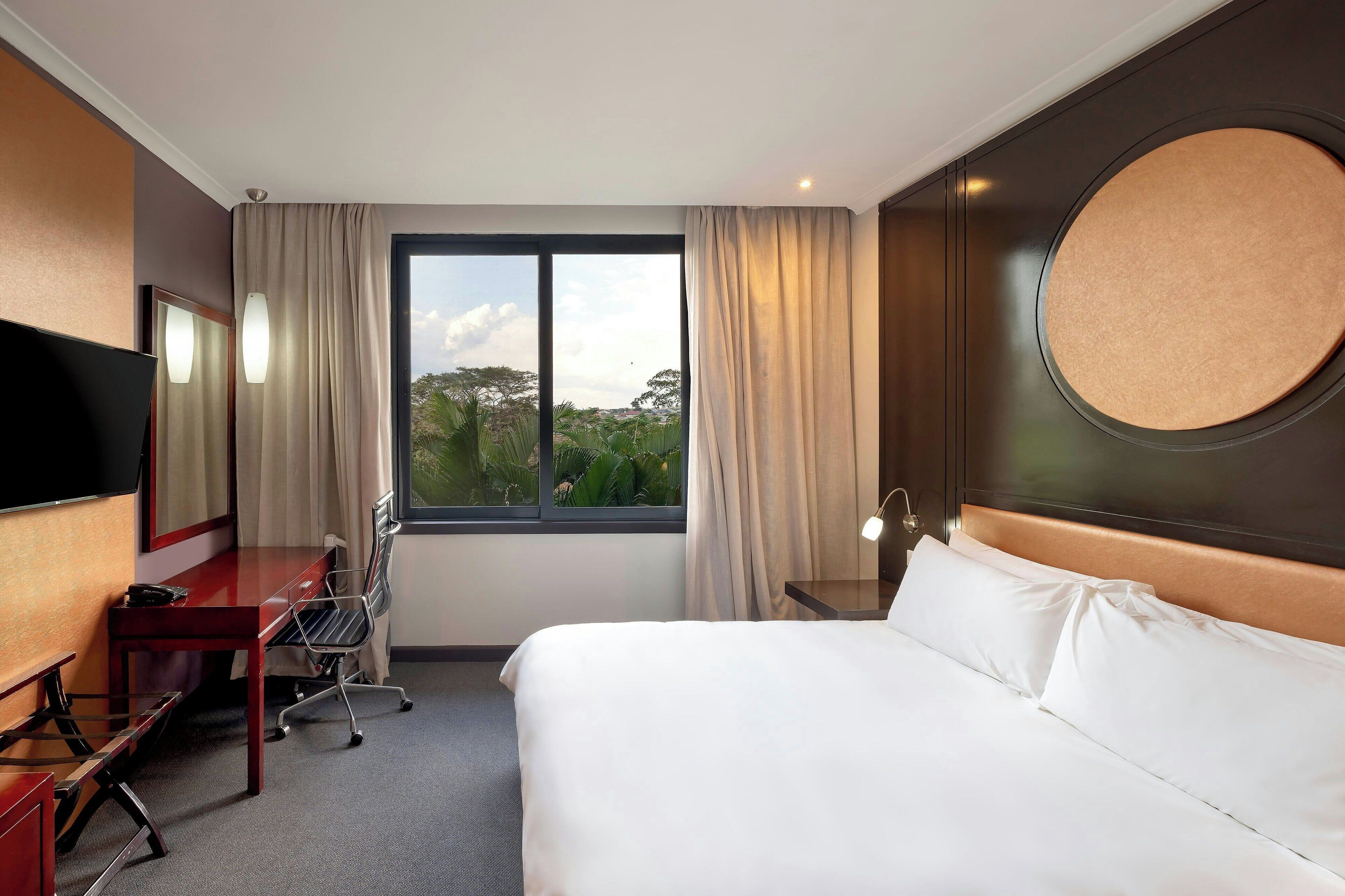 Habitación de hotel con cama, escritorio, silla, televisor, ventana con cortinas y vista a zonas verdes.