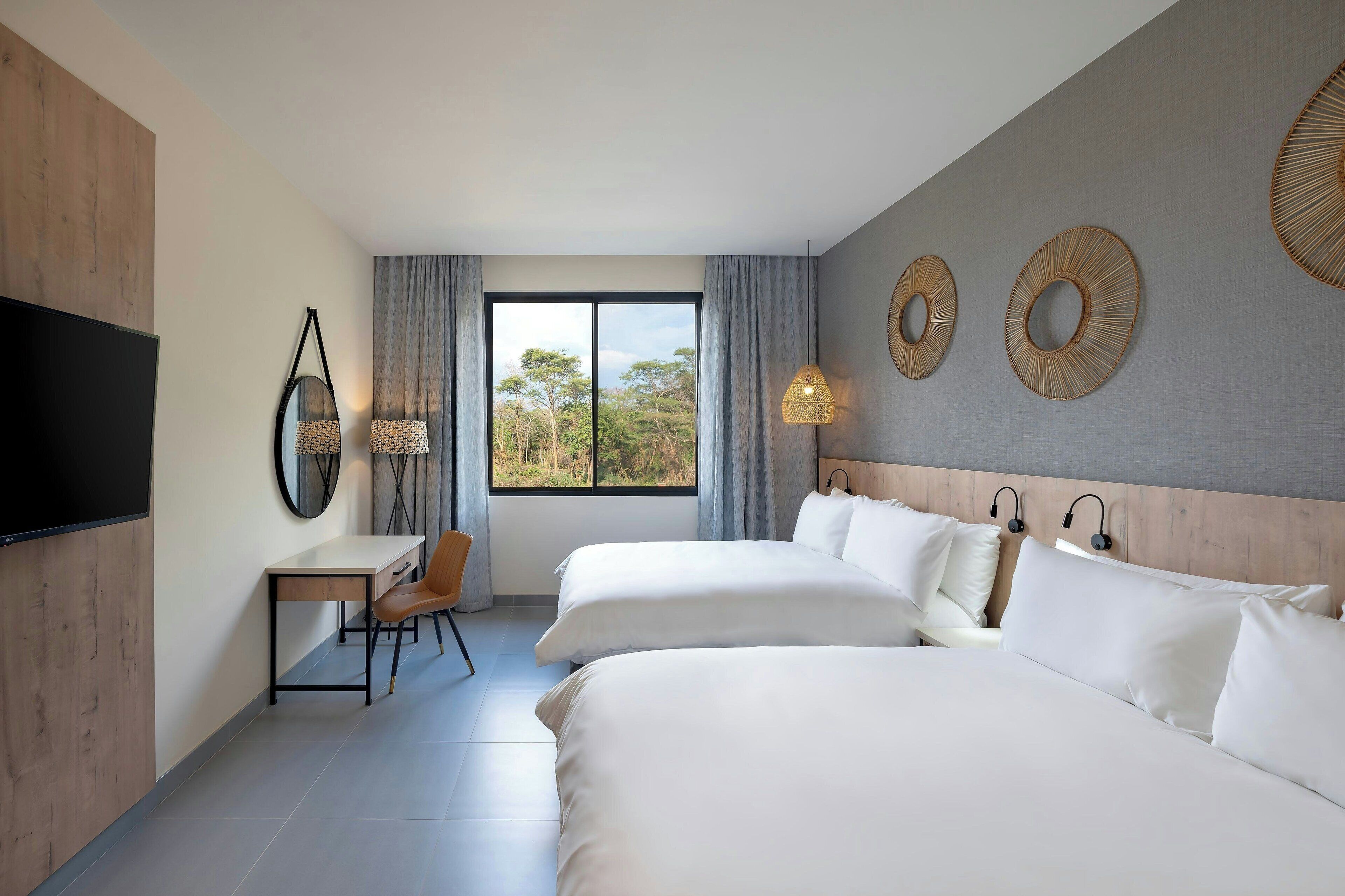 Habitación de hotel con dos camas, un escritorio, una silla, un televisor y un ventanal con vistas a árboles.