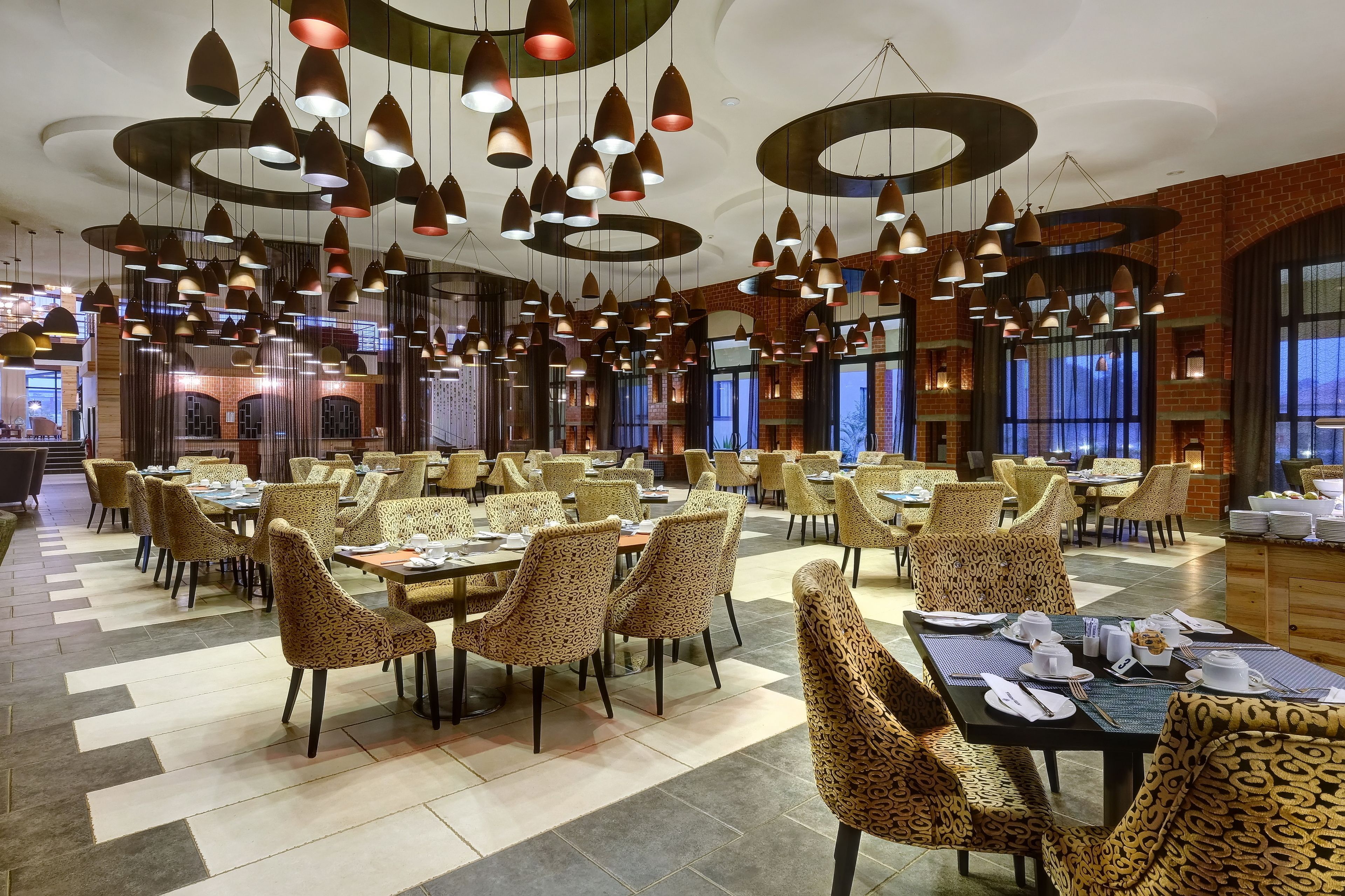 Un vaste espace dédié à la salle à manger, avec de nombreuses tables et chaises, agrémenté d’un lustre au design moderne et de murs en briques apparentes.