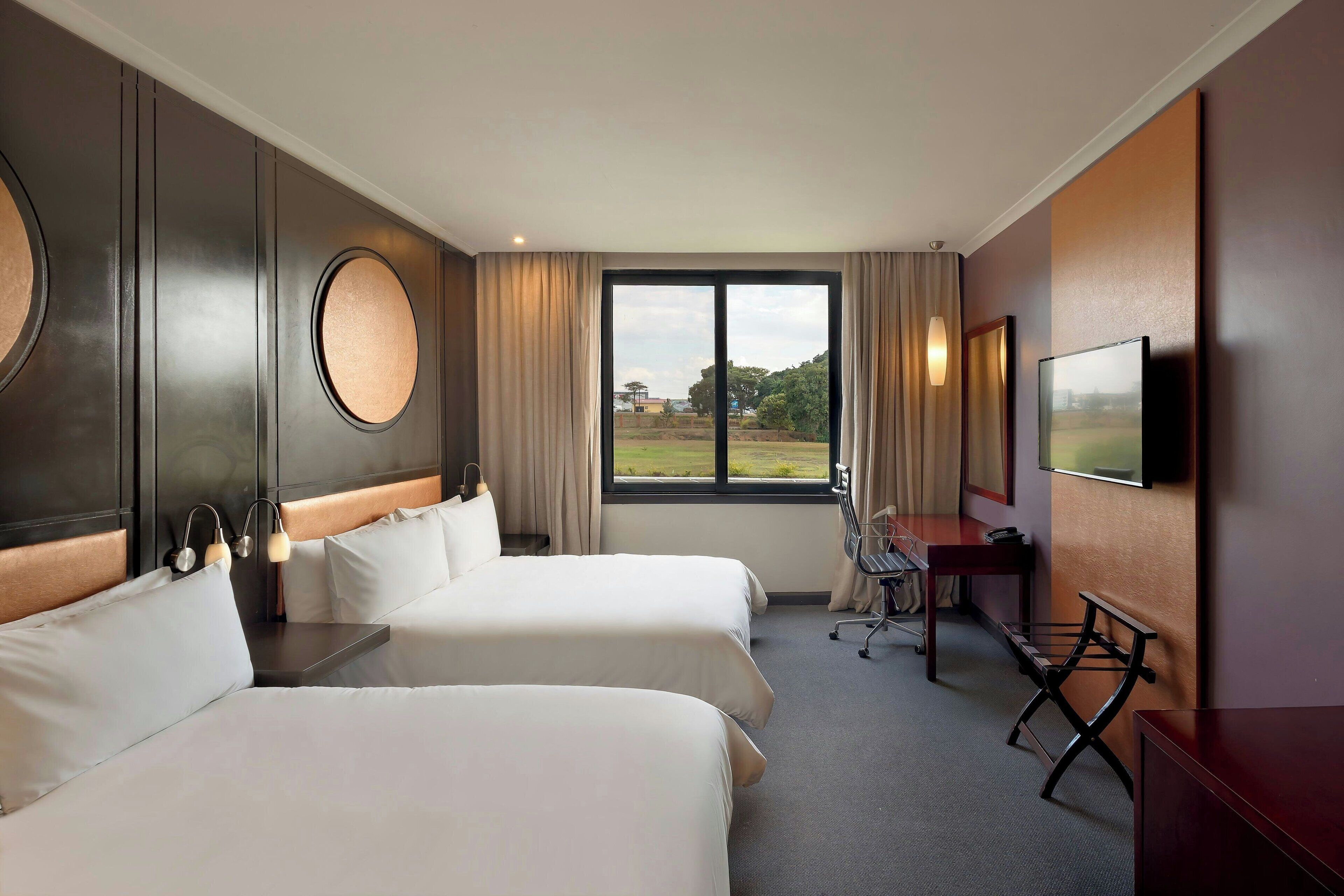 Habitación de hotel con dos camas, un escritorio, una silla, un televisor y una ventana con vistas al exterior.