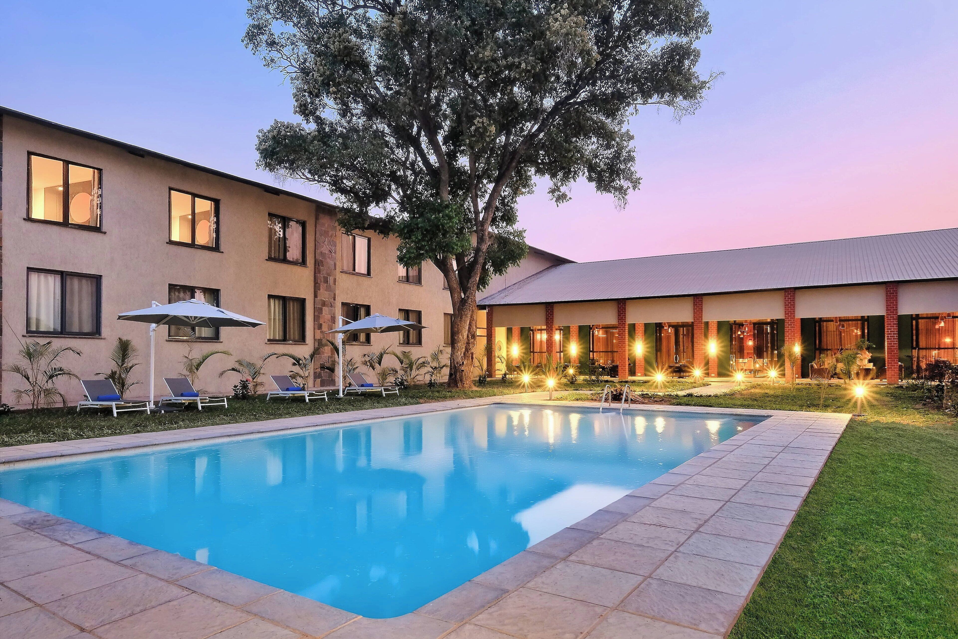 A hotel with a swimming pool, lounge chairs, and a large tree.