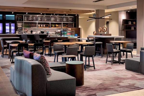 A modern cafe with multiple seating options including tables, chairs, and couches. There is a bar area with a menu board and a coffee machine.
