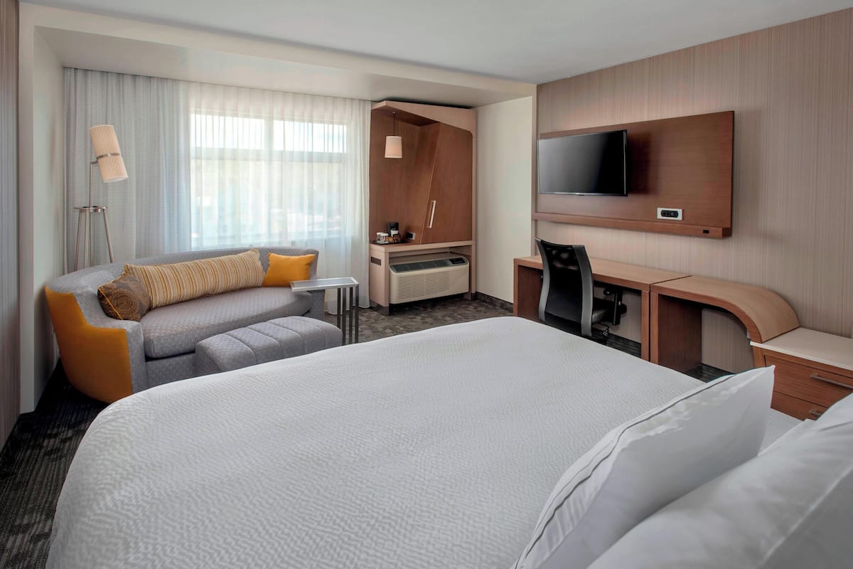 Una habitación de hotel con una cama grande, un escritorio con una silla, un televisor montado en la pared y una zona de estar con sofá y sillón.