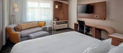 Ein Hotelzimmer mit einem großen Bett, einem Schreibtisch mit Stuhl, einem an der Wand befestigten Fernseher und einer Sitzecke mit Sofa und Sessel.