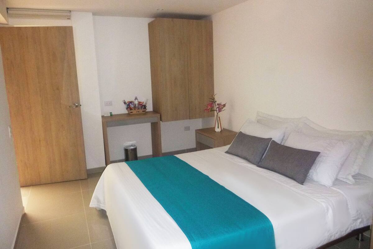 A hotel room with a bed, a nightstand, a small table, a trash bin, and a wooden wardrobe.