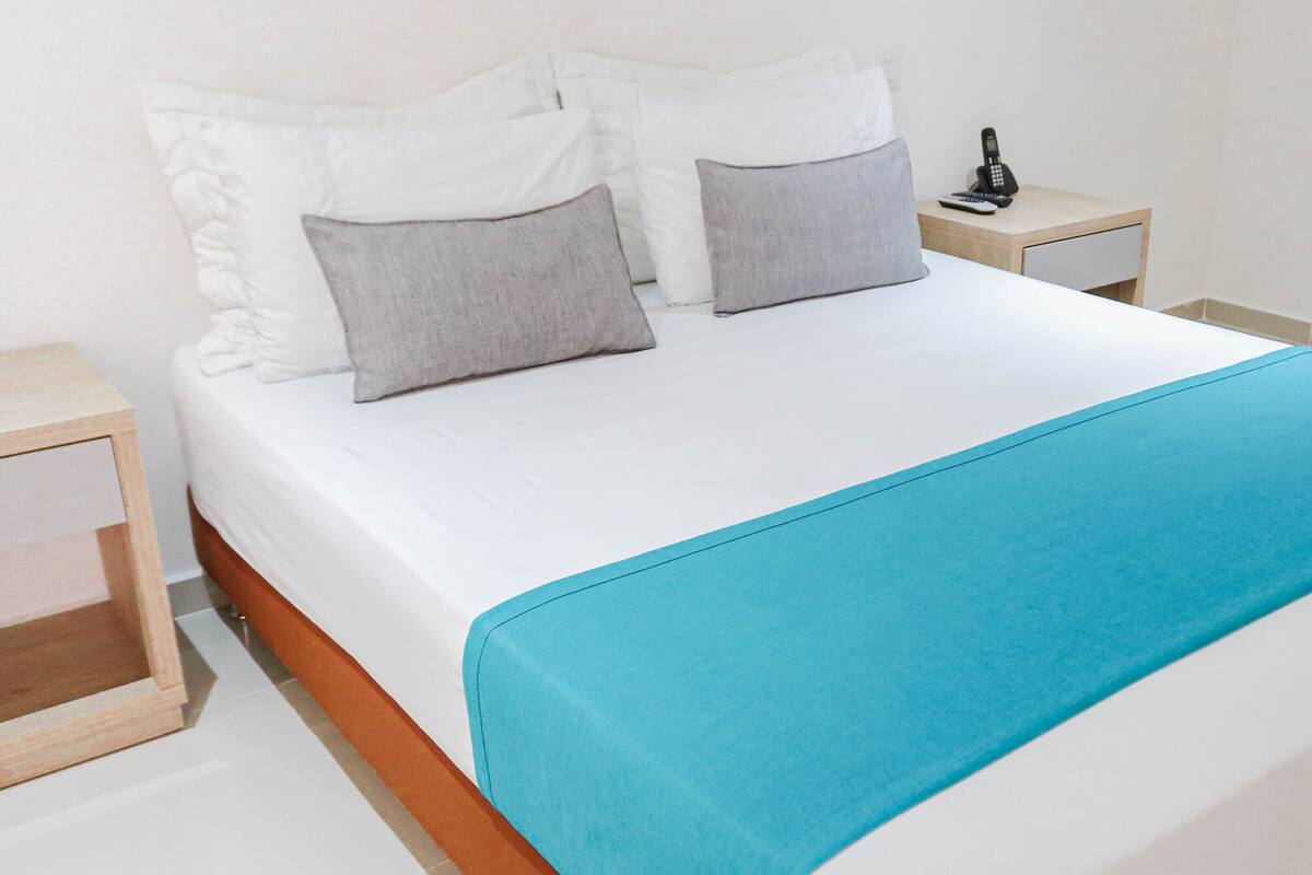 A neatly made bed with white bedding and a blue throw blanket, two gray pillows, and a bedside table with a phone and remote control.