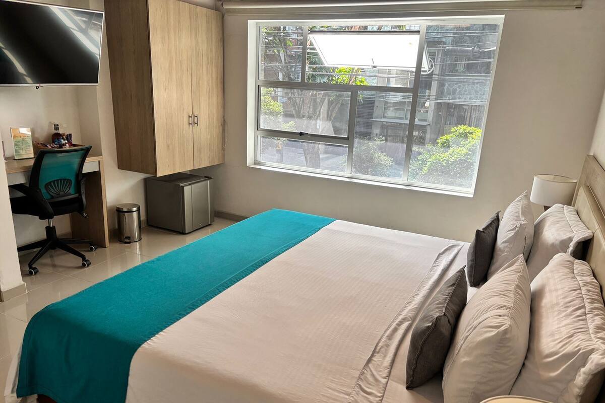A bedroom with a bed, a desk, a chair, a trash bin, and a window with a view of buildings and trees.