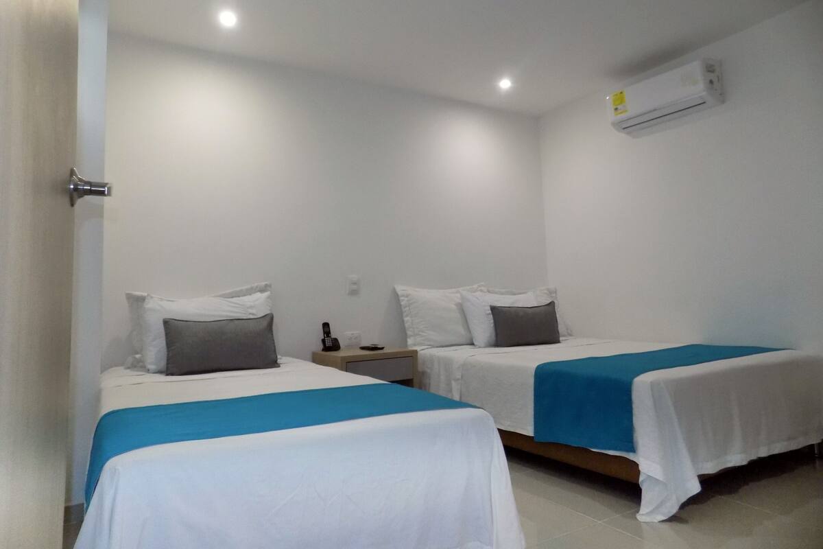 A hotel room with two single beds, each with a blue and white bedspread, a bedside table with a lamp, and an air conditioning unit on the wall.
