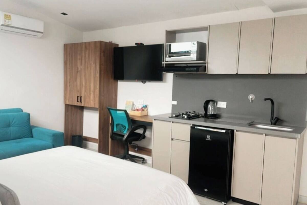 A compact hotel room with a bed, a teal sofa, a wooden wardrobe, a kitchenette with a sink and stove, and a flat-screen TV mounted above the TV.
