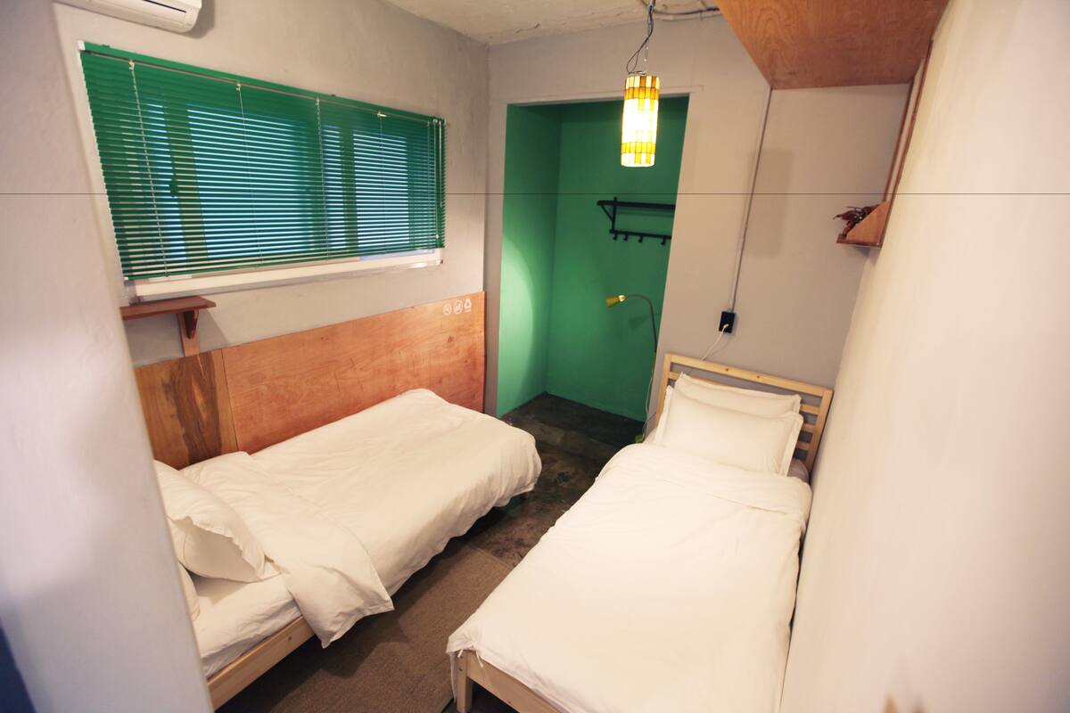 Una habitación pequeña y limpia con dos camas individuales, una puerta verde y una ventana con persianas.