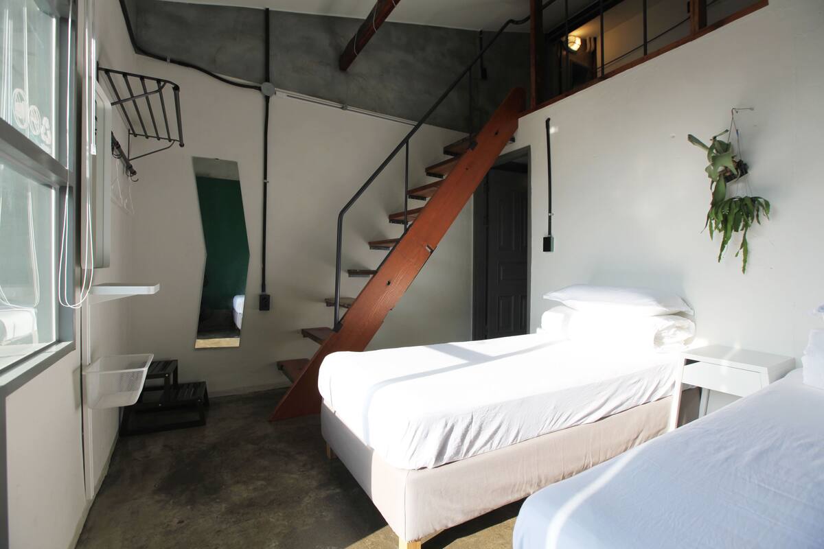 Un dormitorio con cama, una ventana pequeña, una escalera y una planta colgada en la pared.