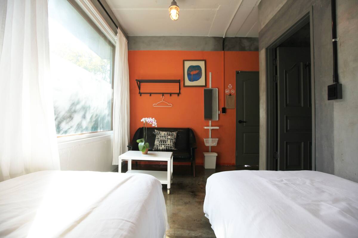 Habitación de hotel con dos camas, una mesita con una planta, un sofá negro y una ventana con cortinas.