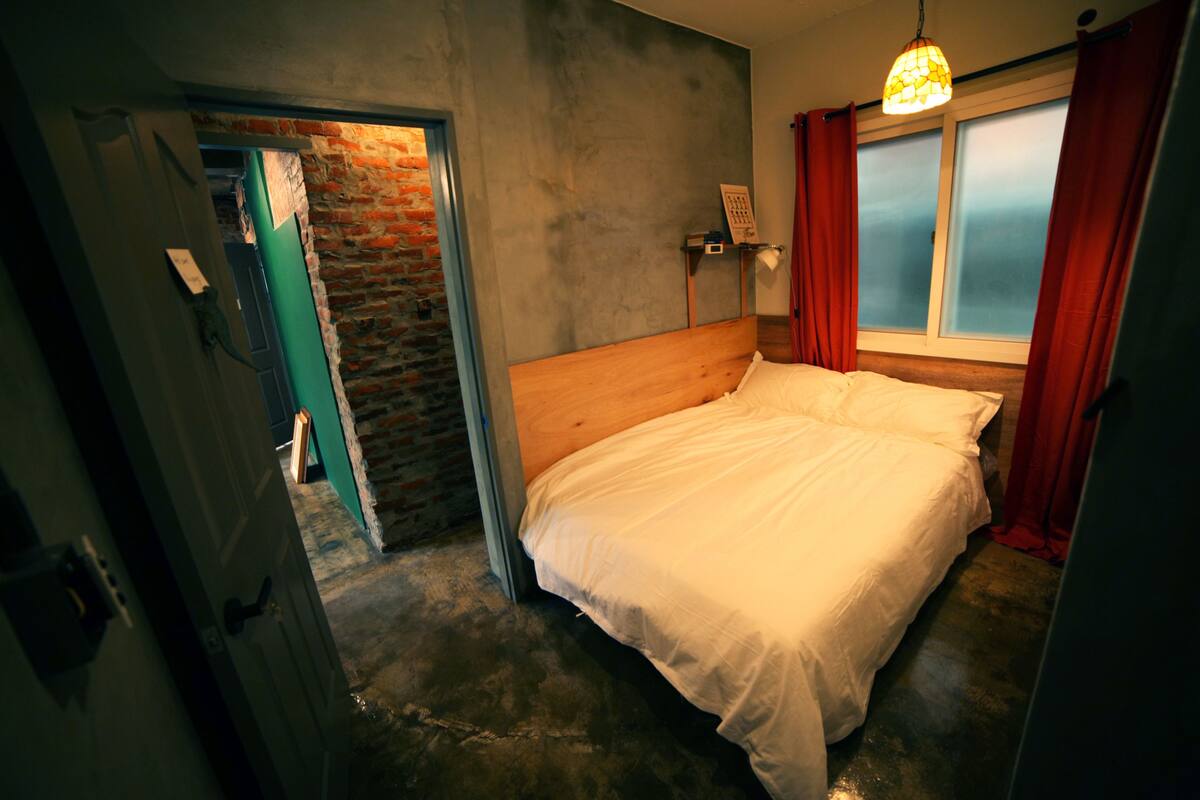 Un dormitorio con cabecera de madera, pared de ladrillo, ventana con cortinas rojas y lámpara colgante.