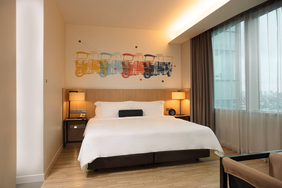 Quarto de hotel com uma cama grande, duas mesas de cabeceira com abajures, vista para a cidade e uma arte vibrante na parede retratando trens.