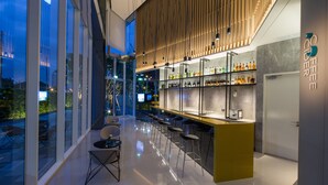 A modern bar area with a long counter, bar stools, and a view of the city through large windows. - Modena by Fraser Bangkok Hotel Residences (Bangkok)