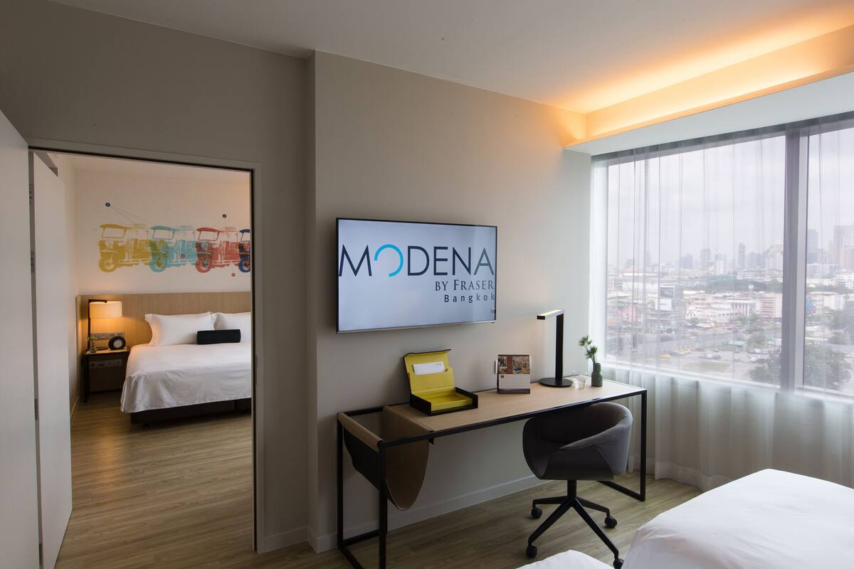 Quarto de hotel moderno com uma cama, escrivaninha, cadeira e uma grande janela com vista para a cidade.