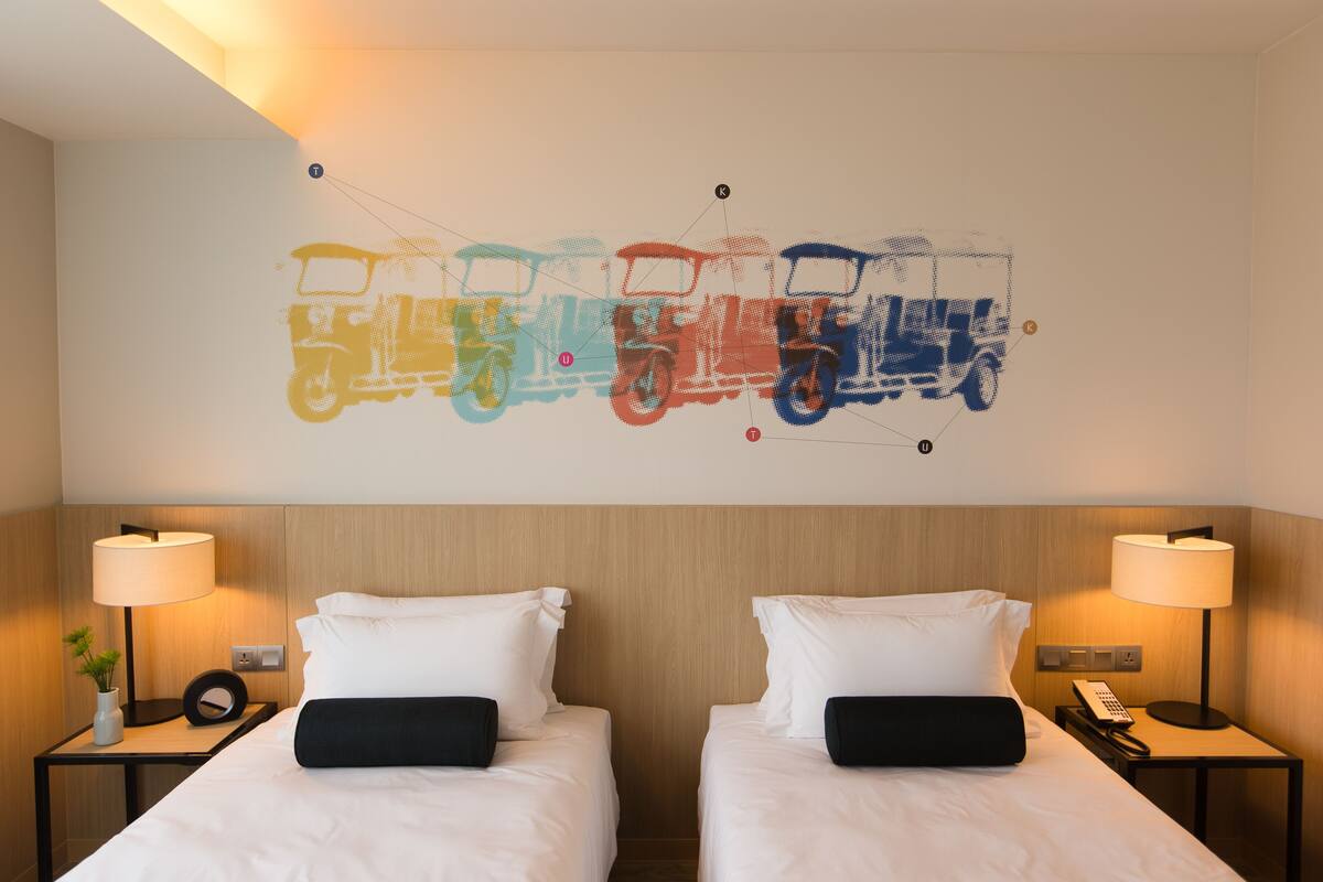 Quarto de hotel com duas camas, uma obra de arte na parede retratando veículos coloridos e luminárias de cabeceira.