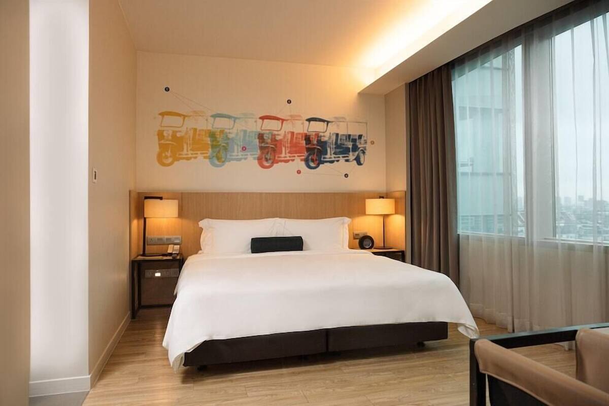 Quarto de hotel com uma cama grande, duas mesas de cabeceira com abajures, uma obra de arte na parede retratando veículos coloridos e uma janela ampla com cortinas.