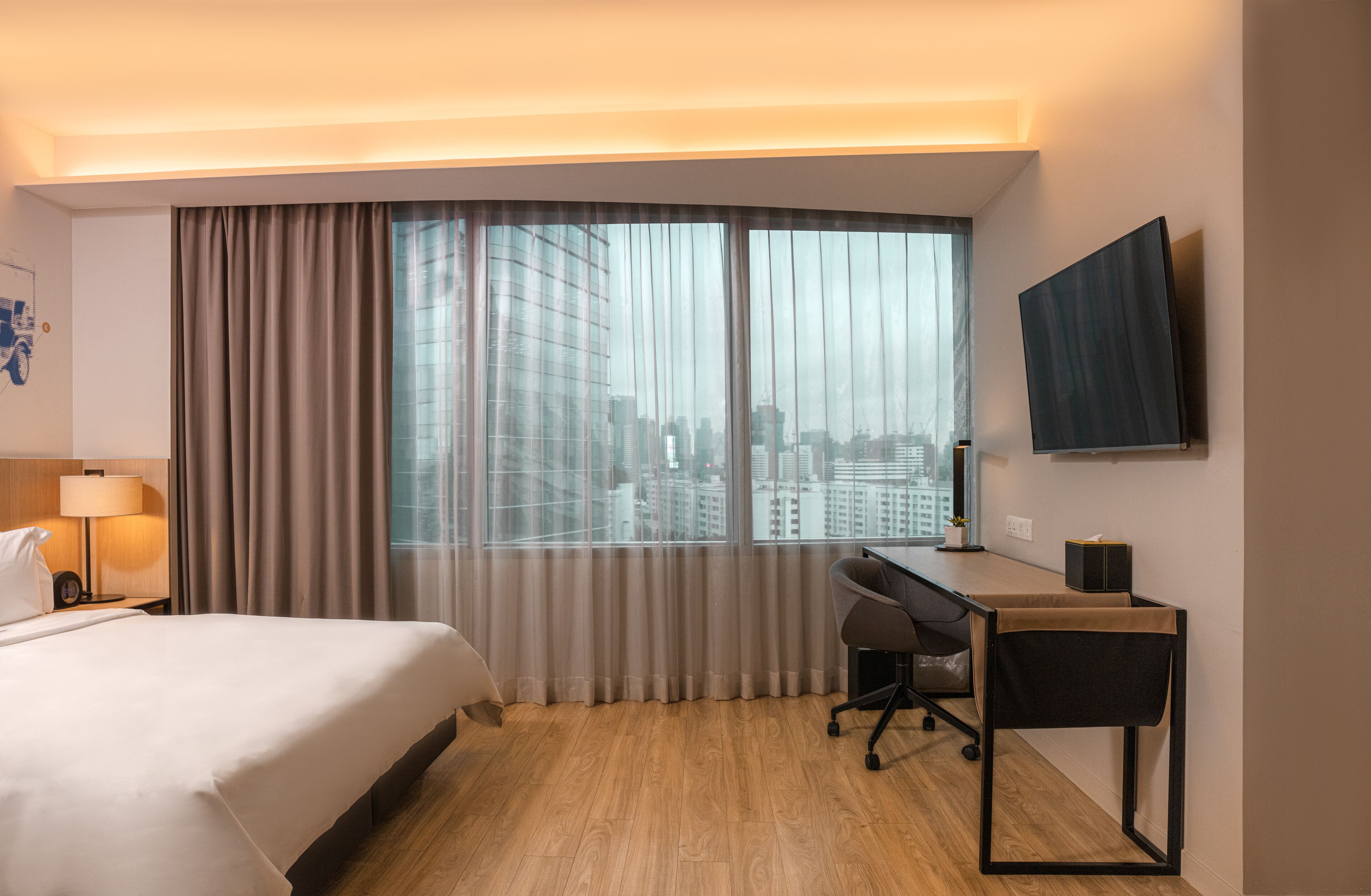 A hotel room with a large bed, a desk, a chair, a TV mounted on the wall, and a view of the city through the window.
