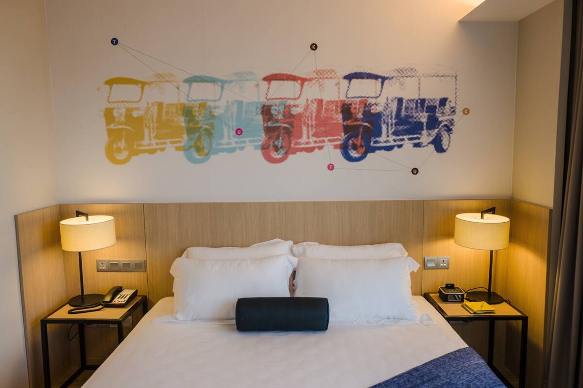 Quarto de hotel com uma cama, duas mesas de cabeceira, uma luminária em cada mesa, uma obra de arte na parede retratando veículos coloridos e uma cabeceira de madeira.