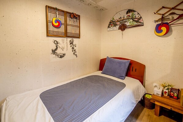 Une chambre à coucher avec un lit, une tête de lit en bois, un ventilateur fixé au mur et une petite table sur laquelle se trouvent un panier et des objets décoratifs.