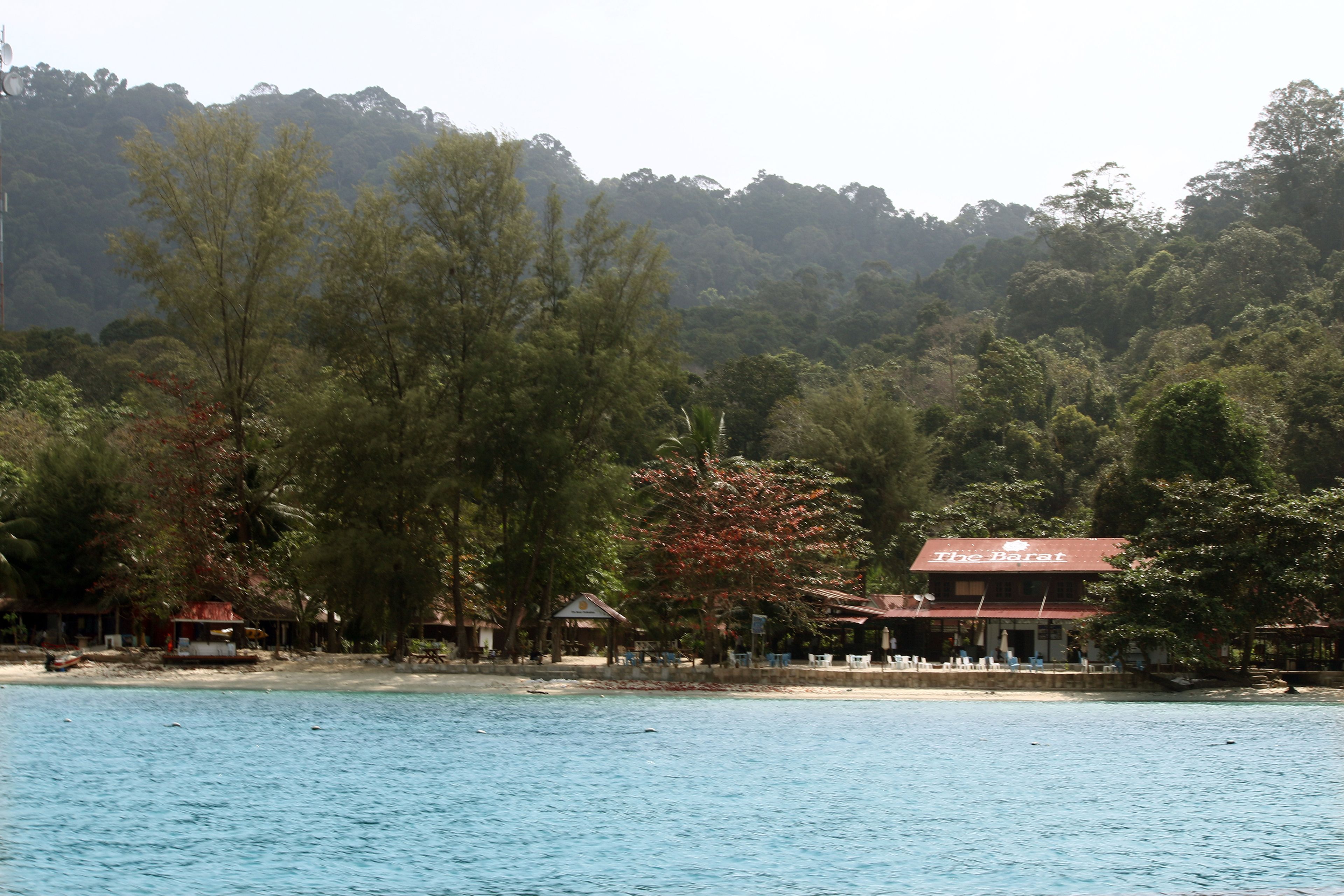A coastal hotel with a red roof, surrounded by dense forest and a beach.