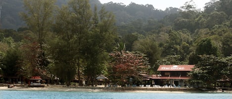 A coastal hotel with a red roof, surrounded by dense forest and a beach.