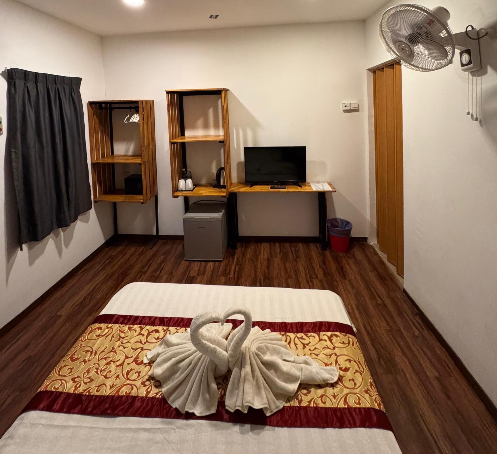 A hotel room with a bed, a desk with a computer, a TV, a fan, and a trash bin.