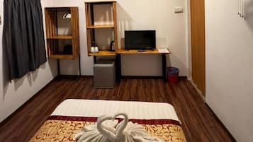 A hotel room with a bed, a desk with a computer, a TV, a fan, and a trash bin.