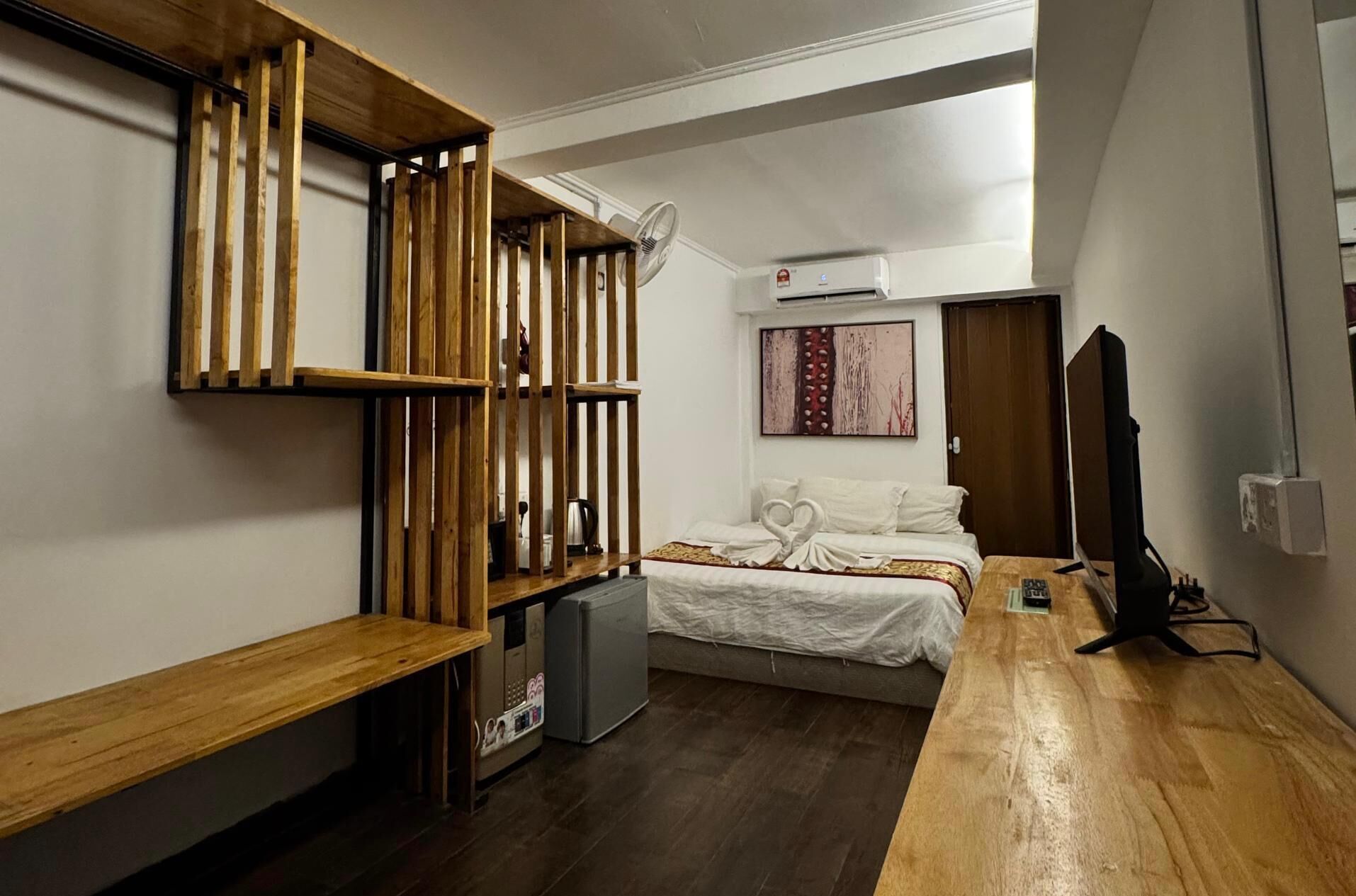 A compact hotel room with a bed, a wooden shelf unit, a small desk, a television, and a kitchenette.