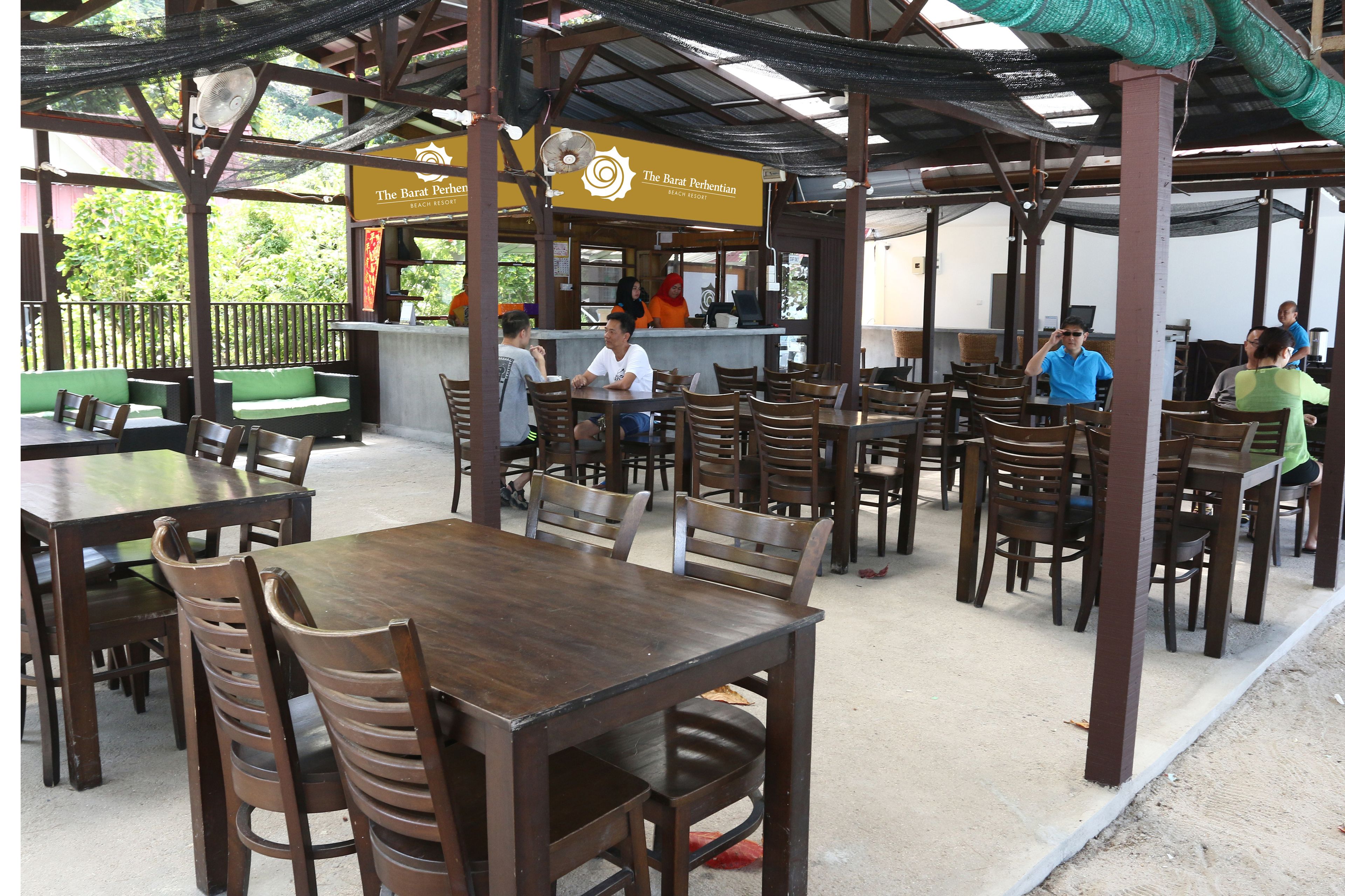 An outdoor restaurant with wooden tables and chairs, a counter with staff, and a sign for 'The Barat Perhentian Beach Resort'.
