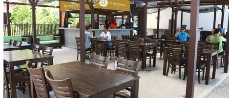 An outdoor restaurant with wooden tables and chairs, a counter with staff, and a sign for 'The Barat Perhentian Beach Resort'.