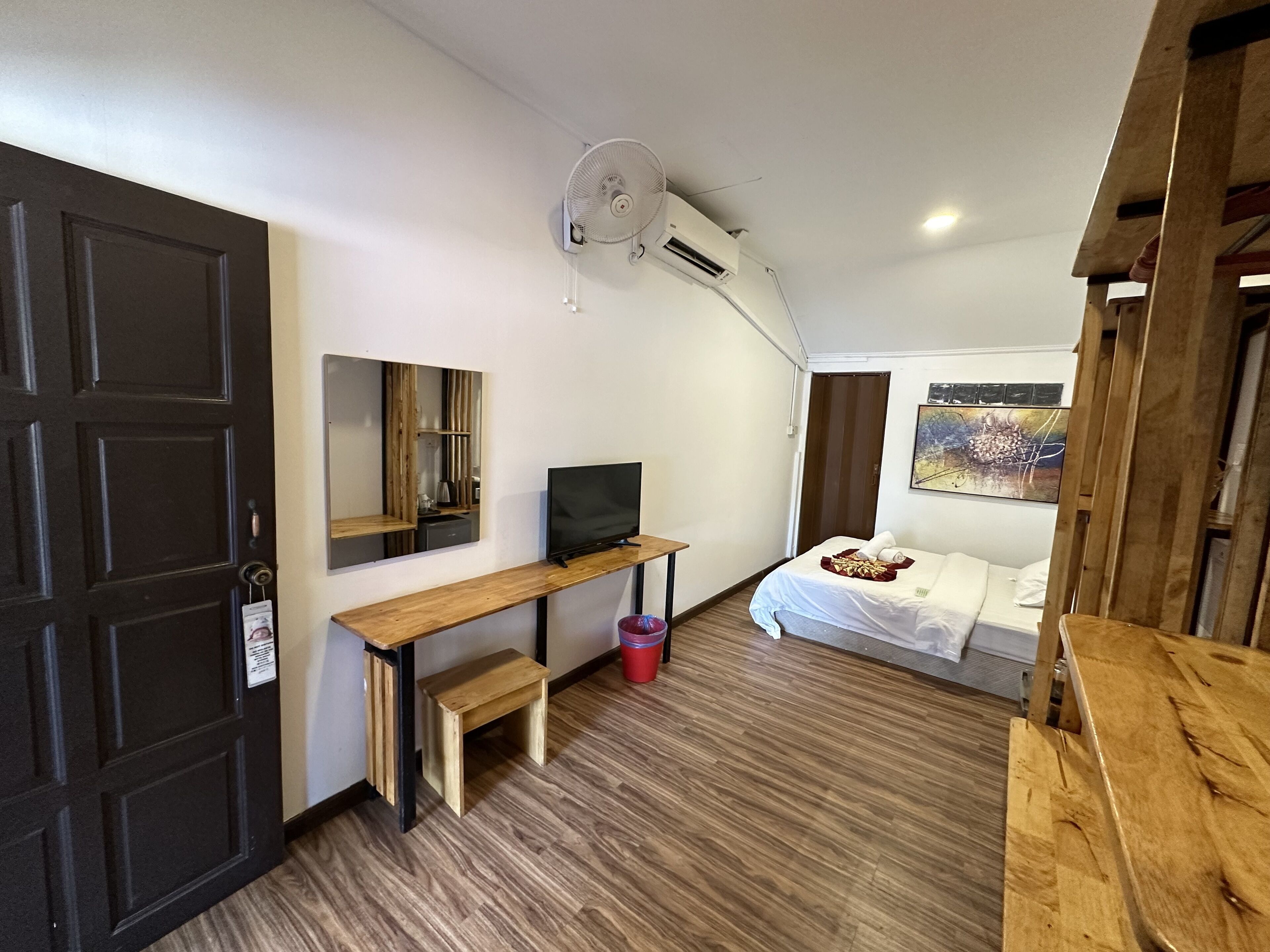 Una habitación pequeña con cama, escritorio, televisor, espejo y ventilador de techo.