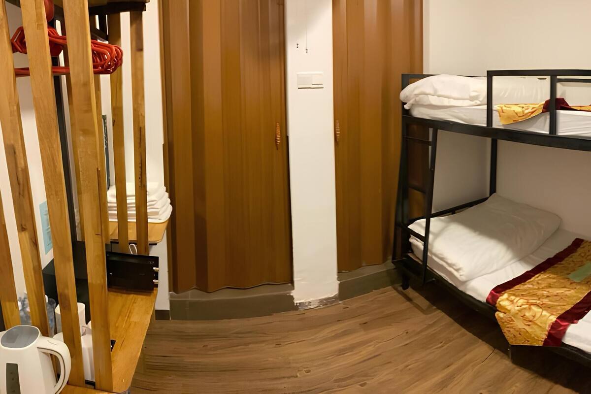 Une chambre avec un lit superposé, un plancher en bois, une bouilloire blanche et un rideau.